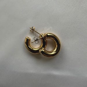 Puede incluir: Un par de pendientes de aro dorados con un dise&ntilde;o grueso y redondeado. Los pendientes est&aacute;n hechos de metal y tienen un acabado pulido.