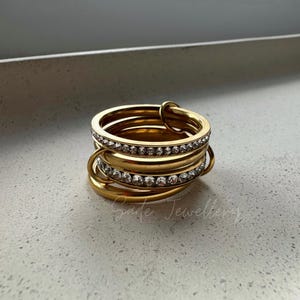 May include: A stackable gold-toned ring set. The rings are connected and feature a row of small, clear crystals. The rings are arranged in a layered design, creating a unique and eye-catching piece of jewelry.