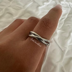 Peut inclure: Une bague argentée avec un design croisé. La bague est lisse et brillante, reflétant la lumière. La bague est portée sur un doigt, avec le texte "Safe Jewellery" visible.