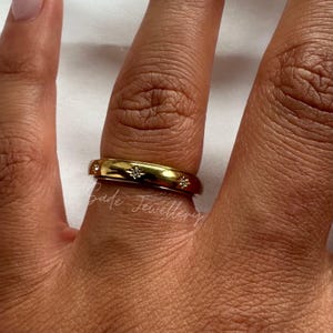 May include: A gold ring with star-shaped detailing is worn on a finger. The ring has a polished surface and is decorated with small, sparkling features. The ring is a piece of jewellery.