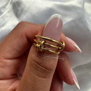 Könnte beinhalten: Goldring mit einem geknoteten Design. Der Ring besteht aus mehreren Bändern aus Golddraht, die miteinander verdreht sind, um einen Knoten zu bilden.