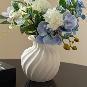 Peut inclure: Un vase en céramique blanche avec un motif en spirale contient un bouquet de fleurs artificielles. Les fleurs comprennent des hortensias blancs, des orchidées bleues et des clématites violettes.