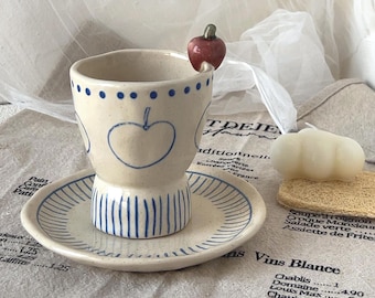 May include: A cream-colored ceramic cup and saucer set featuring hand-painted blue apple outlines and vertical stripes. The cup has a small, sculpted red apple attached to the rim. The design includes a row of blue dots around the upper edge.