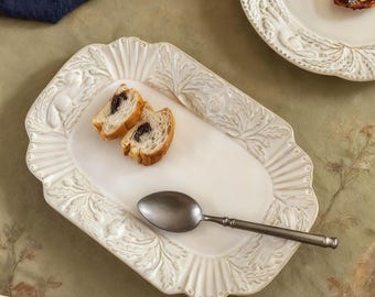 May include: A white, rectangular serving platter with a floral design and scalloped edges. It holds two slices of bread and a silver spoon. A white plate with food and a wooden pepper grinder are also visible.