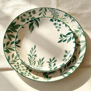May include: A set of two round ceramic plates featuring a cream-coloured base with a painted pattern of dark green leaves and delicate foliage. The larger plate sits beneath the smaller plate, both displaying consistent botanical illustrations.