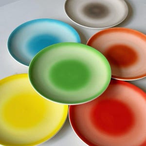 May include: A collection of six round ceramic plates featuring a gradient color design. The set includes individual plates in solid shades of bright yellow, lime green, sky blue, soft orange, deep red, and a neutral greyish-beige tone.