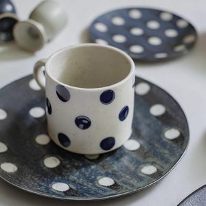 Può includere: Un set di tazza e piattino in ceramica color crema, cosparso di pois blu navy. La tazza ha pois e un manico, ed è appoggiata su un piatto abbinato con pois bianchi. Altri piatti e tazze abbinati sono visibili sullo sfondo.