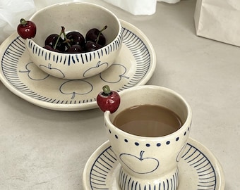 May include: A ceramic set featuring a bowl and a cup, each with a matching saucer. The off-white pieces are decorated with blue hand-painted apple outlines and striped rims. A small, three-dimensional red cherry accent is attached to the rim of both the bowl and the cup, which contains a brown liquid.