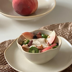 May include: A white ceramic bowl filled with a creamy yogurt parfait topped with sliced peaches, kiwi, almonds, and green jalapeno peppers. The bowl is sitting on a white ceramic plate.