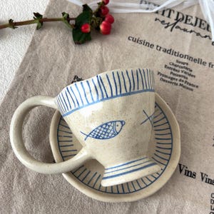 Puede incluir: Un juego de taza y platillo de cerámica con diseño de pez. La taza y el platillo son de color blanquecino con rayas azules y una ilustración de pez. El juego está sobre una superficie beige con texto.