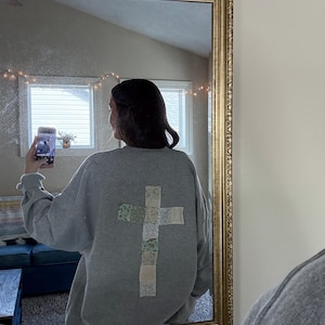 May include: A gray sweatshirt with a patchwork cross on the back. The cross is made of different floral fabrics in shades of green, white, and pink.