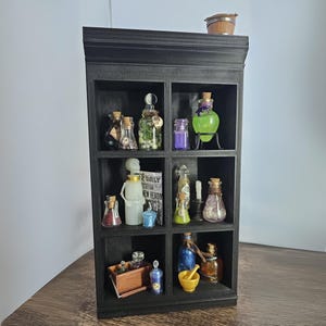 Puede incluir: Una estantería de madera negra con nueve compartimentos, cada uno lleno de varias botellas y frascos de vidrio que contienen líquidos y objetos coloridos. La estantería tiene una pequeña caja de madera y un mortero y un pilón en el estante inferior. La estantería está decorada con un pequeño cubo de madera en la parte superior.