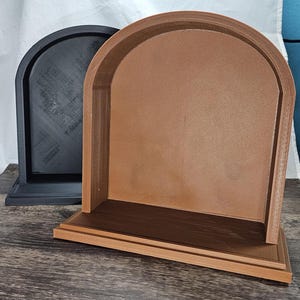 May include: Two arched display shelves, one black and one brown, stand side-by-side. The shelves have a rectangular base and a curved top, creating a niche-like space. The brown shelf has a darker brown interior.