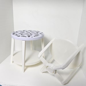 May include: A small, white, round table with a patterned top featuring black feather designs. The table has four legs and a lower shelf. A white folding chair is next to the table.