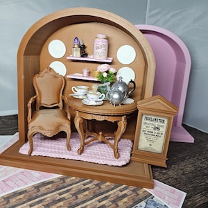 Könnte beinhalten: Miniatur-Teeservice-Szene mit kleinem Holztisch, Stuhl und silberner Teekanne. Die Szene umfasst Teetassen, einen rosa Teppich und ein Schild mit der Aufschrift "Proclamation". Der Hintergrund ist ein brauner Bogen mit Regalen.