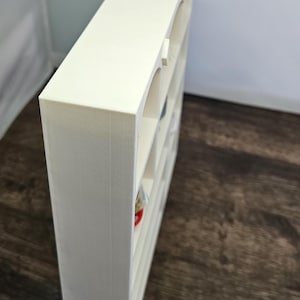 May include: A cream-colored, rectangular shelf unit with multiple compartments. The shelf has an arched design on the front of each compartment. The unit is made of a solid material and is positioned at an angle, showcasing its depth and structure.