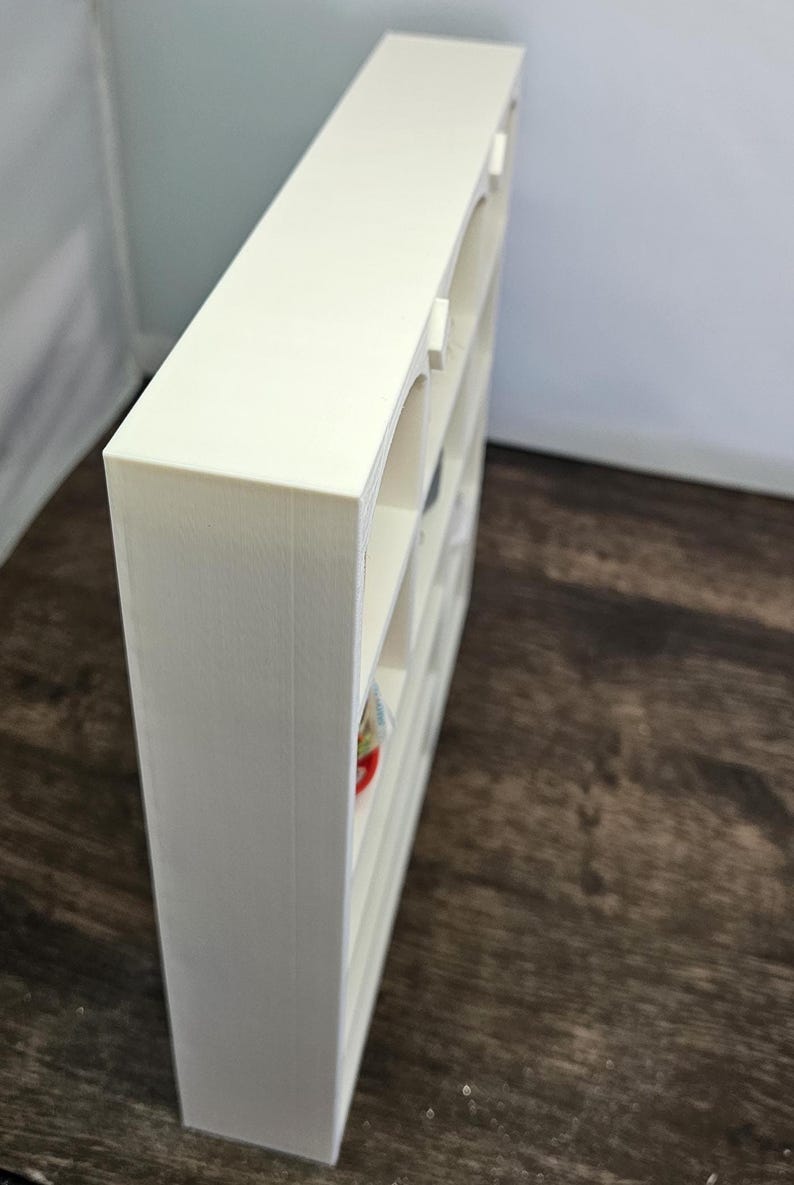 May include: A cream-colored, rectangular shelf unit with multiple compartments. The shelf has an arched design on the front of each compartment. The unit is made of a solid material and is positioned at an angle, showcasing its depth and structure.