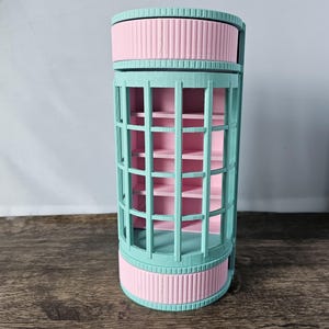 May include: A cylindrical storage container with a pastel color scheme. The container features a light teal cage-like exterior, pink interior shelves, and pink and teal ribbed bands at the top and bottom. The design is reminiscent of a decorative birdcage.