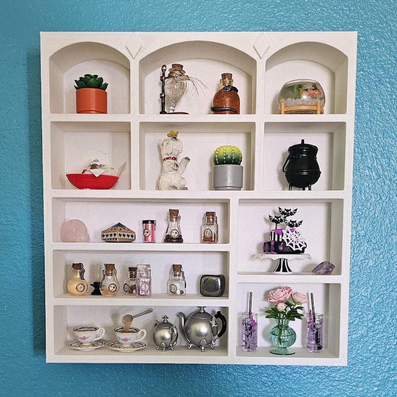 White mini display shelf unit with 12 compartments, featuring various decorative items, including a small potted plant, a miniature cauldron, a small cake, and several glass bottles.