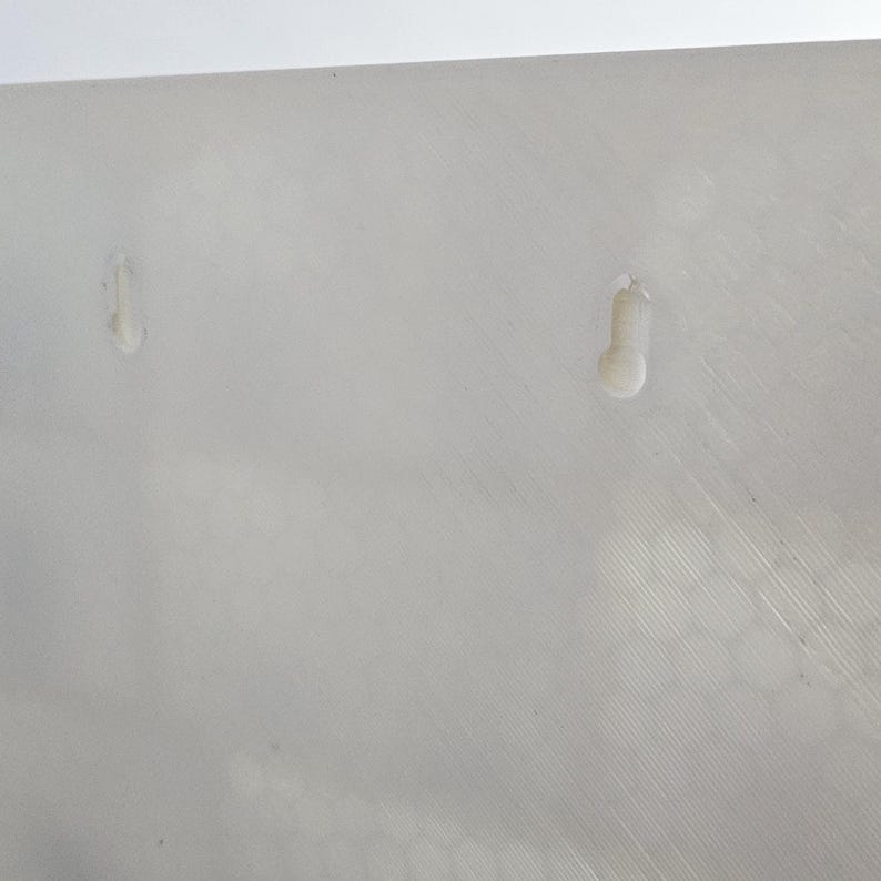 May include: Close-up of a white, textured surface with two small, raised, teardrop-shaped protrusions. The surface has subtle diagonal lines and a slightly uneven texture, suggesting a manufactured material.