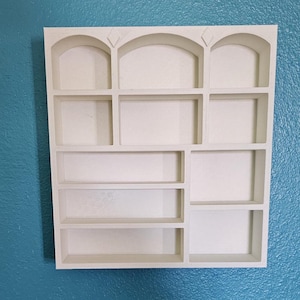May include: A white, multi-compartment wall shelf with a variety of rectangular and arched openings. The shelf is mounted on a textured, teal-colored wall. The top row features three arched openings with diamond-shaped accents.