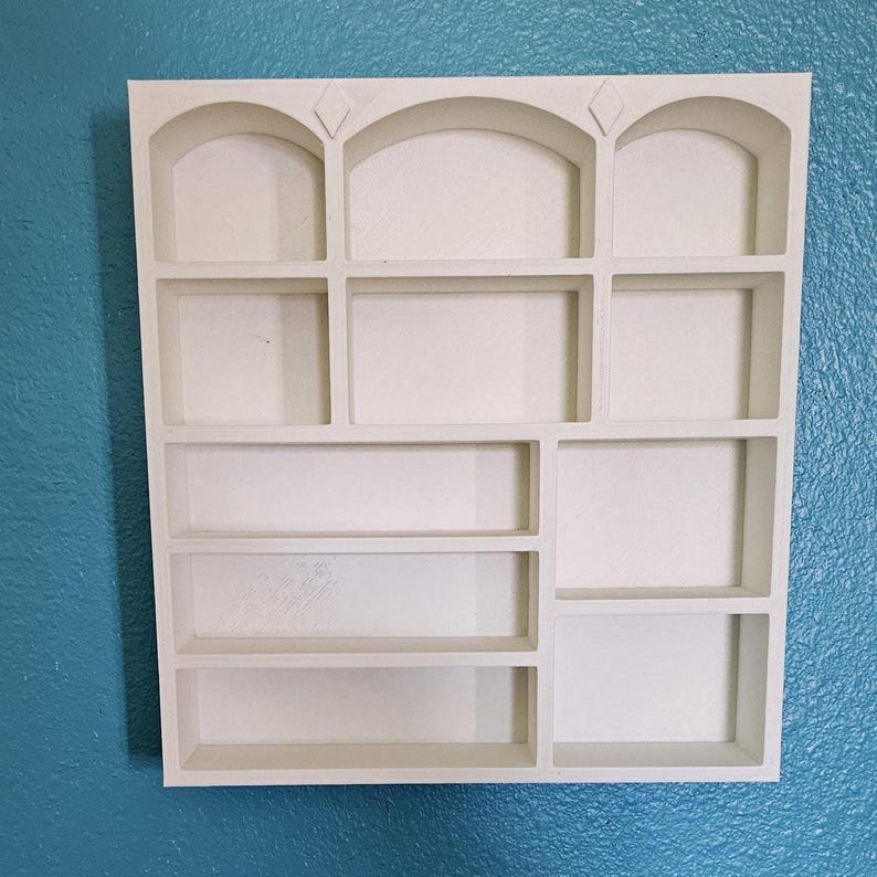 May include: A white, multi-compartment wall shelf with a variety of rectangular and arched openings. The shelf is mounted on a textured, teal-colored wall. The top row features three arched openings with diamond-shaped accents.