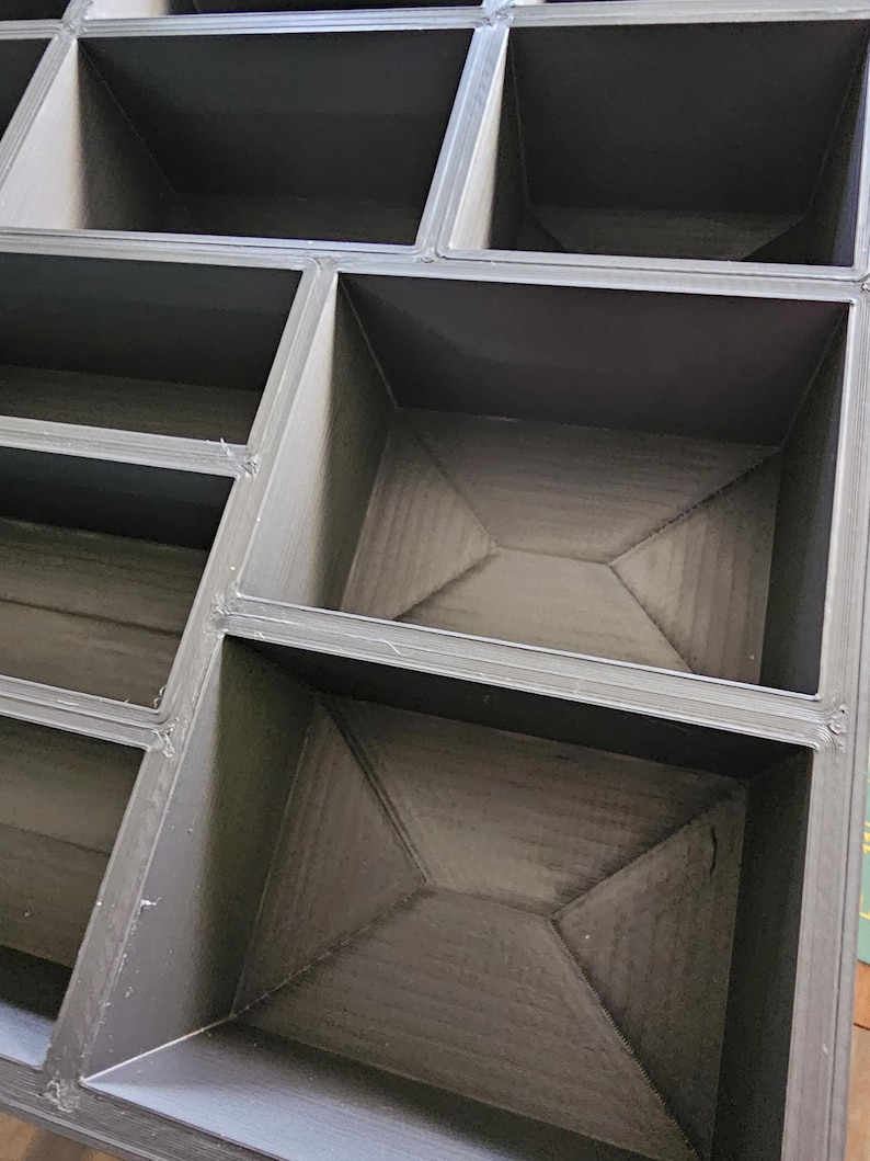 K&ouml;nnte beinhalten: Ein modulares, dunkelgraues Aufbewahrungselement mit mehreren F&auml;chern. Das Element ist mit geraden Linien und rechten Winkeln konstruiert, wodurch ein geometrisches Muster entsteht. Das Innere jedes Fachs hat ein strukturiertes Aussehen.