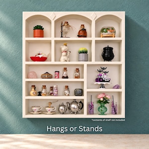 May include: A cream-colored wooden display shelf featuring multiple arched compartments and rectangular cubbies. The unit is designed to hang on a wall or stand on a flat surface. The text "*contents of shelf not included" and "Hangs or Stands" is visible.