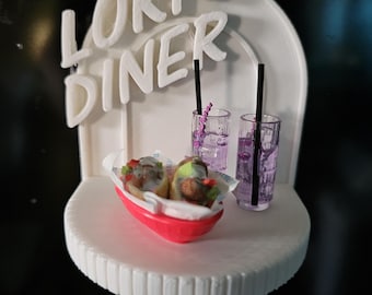 Puede incluir: Un modelo 3D impreso en blanco de un restaurante con las palabras "Lori's Diner" en la parte delantera. El modelo incluye dos vasos de líquido morado con pajitas y una bandeja roja con dos sándwiches.