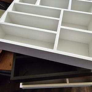 Puede incluir: Una estantería de pared de madera blanca con múltiples compartimentos de varios tamaños. La estantería tiene forma rectangular con un borde gris y está diseñada para exhibir objetos pequeños o coleccionables. La estantería está vacía.