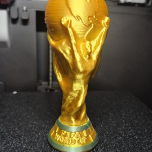 May include: A gold-colored trophy with a globe on top, held by two figures. The base of the trophy reads "FIFA WORLD CUP" in green lettering. The trophy is likely a replica or a decorative item.