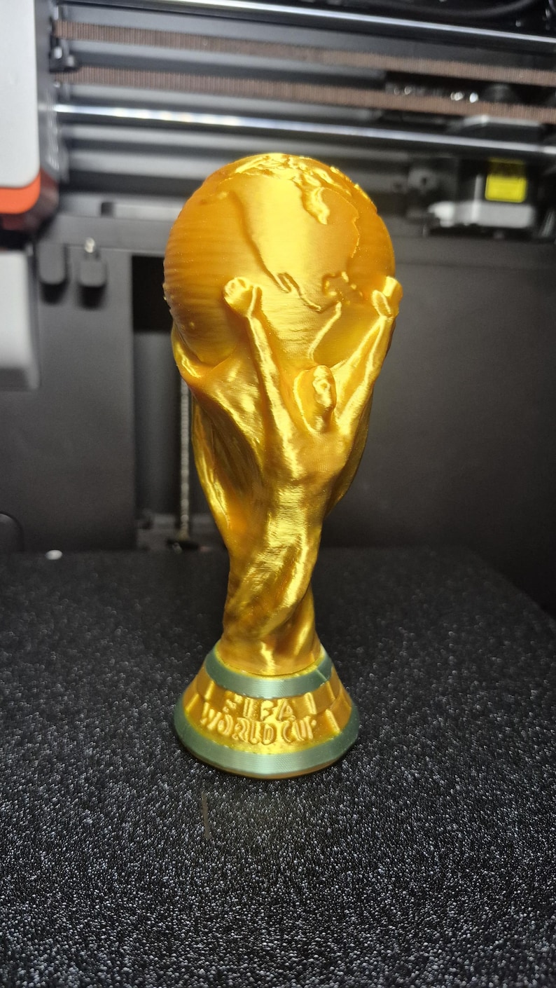 May include: A gold-colored trophy with a globe on top, held by two figures. The base of the trophy reads "FIFA WORLD CUP" in green lettering. The trophy is likely a replica or a decorative item.