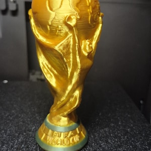 May include: A gold-colored trophy with a globe on top and the words "FIFA WORLD CUP" on the base. The trophy is made of a shiny material and is set against a dark background.