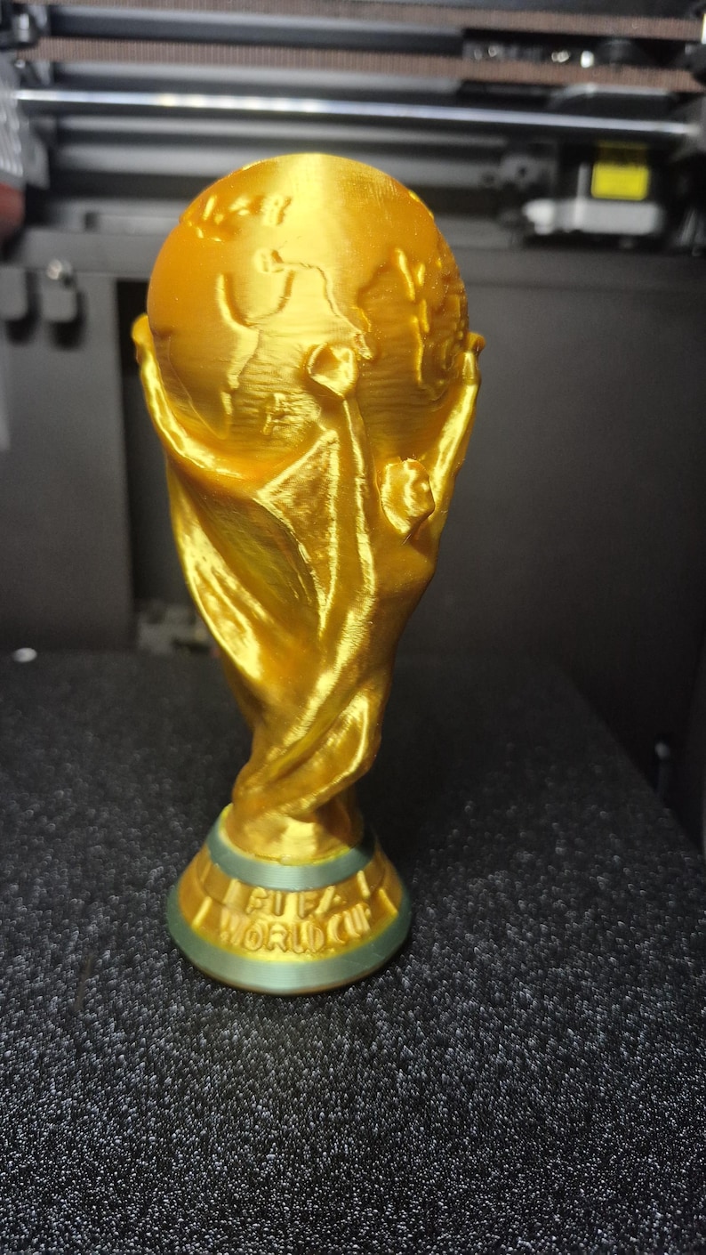 May include: A gold-colored trophy with a globe on top and the words "FIFA WORLD CUP" on the base. The trophy is made of a shiny material and is set against a dark background.
