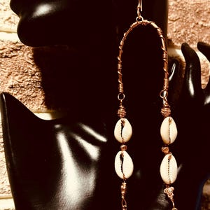 May include: Copper-toned wire earrings featuring a curved design. Each earring has three white seashells and gold-colored accents. The earrings dangle from a hook and end with a small gold-colored bell.