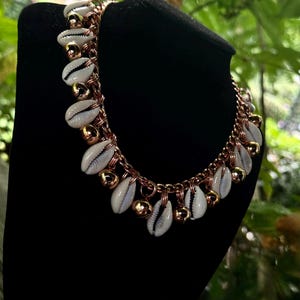 May include: A necklace featuring white cowrie shells and gold-colored beads strung on a copper-toned chain. The necklace is displayed on a black velvet bust, with green foliage in the background. The jewelry has a bohemian style.