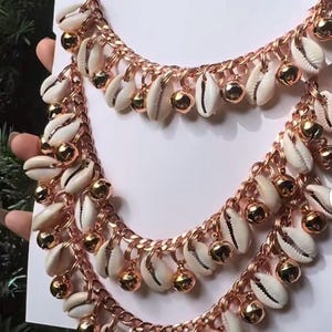 May include: Two layered necklaces with a rose gold chain, adorned with white seashells and small gold-colored bells. The text "The originals" is at the top of the image. The jewelry is displayed on a white background.