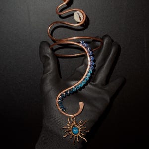 Può includere: Bracciale in filo di rame a forma di serpente, ornato di perline blu e turchesi. Il bracciale presenta un ciondolo a forma di sole con un centro blu. Un cristallo blu si trova sulla sommità della testa del serpente. Il bracciale è tenuto da una mano guantata.
