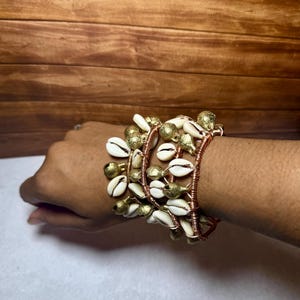 May include: A multi-strand bracelet constructed from copper-colored wire wrapped around the wrist, featuring an alternating arrangement of small, polished gold-toned metal bells and white, oval-shaped cowrie shells throughout the design.