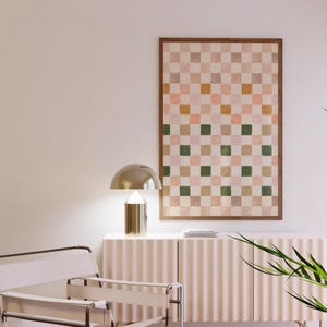 May include: A white chair with a metal frame sits in front of a light pink cabinet with a gold lamp on top. A framed print with a checkered pattern in shades of pink, green, and brown hangs on the wall behind the cabinet.