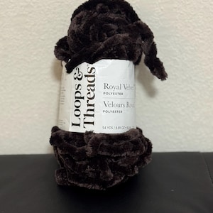 Può includere: Una bobina di filato Royal Velvet in poliestere marrone scuro di Loops & Threads. L'etichetta indica 49.38 metri e 251.7 grammi di filato. Il filato è un materiale morbido e felpato, ideale per progetti di maglia e uncinetto.