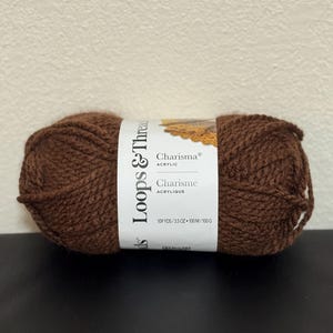 Puede incluir: Ovillo de hilo acrílico marrón de Loops & Threads. La etiqueta dice "Charisma" y "Charisme" con medidas de 100 metros/100g. El hilo es de color marrón oscuro liso.