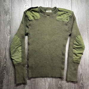 May include: An olive green ribbed knit sweater with reinforced patches on the shoulders and elbows. The long-sleeved sweater has a crew neck and is displayed on a wooden surface.