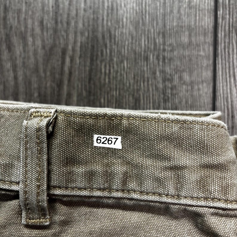 Puede incluir: Primer plano de tela verde oliva con trabilla y costuras visibles. Una peque&ntilde;a etiqueta blanca con el n&uacute;mero "6267" est&aacute; adherida a la tela. El fondo es una superficie de madera oscura y texturizada.