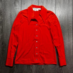 Può includere: Una camicia rossa a maniche lunghe con abbottonatura, un ampio colletto a punta e polsini larghi. La camicia presenta una fila verticale centrale di piccoli bottoni e un'etichetta interna bianca sul collo con il testo Sport Couture by Christian Dior.