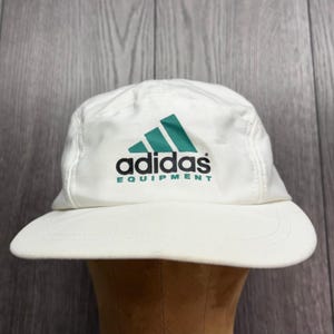 May include: A white five-panel baseball cap featuring an embroidered green Adidas logo above the black text adidas and the green text EQUIPMENT. The cap has a flat brim and a structured crown, showcasing a clean design with bold branding.