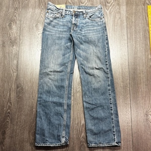 Könnte beinhalten: Hellblaue Jeans mit Knopfleiste und klassischem Fünf-Taschen-Design. Die Jeans haben eine verwaschene Waschung und subtile Abnutzungserscheinungen, die ihnen einen Used-Look verleihen. Die Jeans liegen flach auf einer Holzoberfläche.