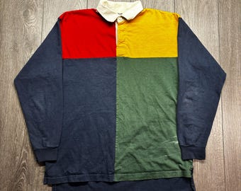 Puede incluir: Una camiseta de rugby de manga larga con un diseño de bloques de color. La camiseta presenta un cuello blanco y una parte delantera dividida en cuatro secciones: rojo, amarillo, verde y azul marino. Las mangas son azul marino.