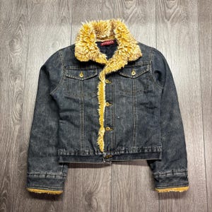 May include: A dark wash denim jacket with a faux fur collar and trim. The jacket has gold-colored buttons and pockets on the chest. The label inside the collar reads "NEVADA".