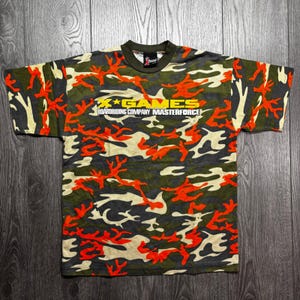 May include: Camouflage pattern t-shirt with the text "X Games Boardriding Company Masterforce" in yellow and white. The shirt features a mix of green, orange, and cream colors, creating a military-inspired design. The shirt has a crew neck and short sleeves.
